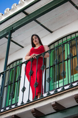 A beautiful teenage girl with long dark hair, dressed in red and wearing high heels, leaning on a staircase railing, with a wooden door in the background. Photo taken from below.