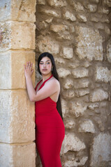 A beautiful teenage girl with long dark hair, dressed in red and wearing high heels, leaning under a Roman archway on a street in a typical Andalusian town. In the background is a wall.