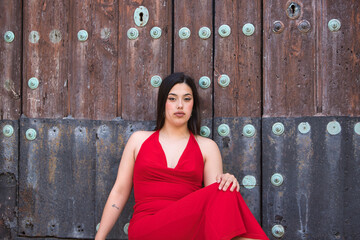 A pretty teenage girl with long dark hair, dressed in red and wearing high heels, sitting on the steps of a church and leaning against the wooden door.