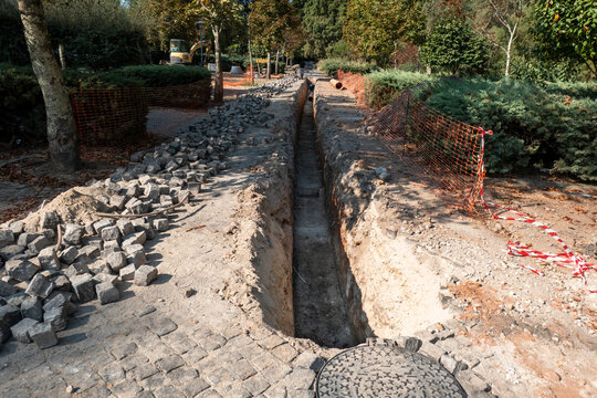 Obra de engenharia civil para instala&ccedil;&atilde;o de saneamento p&uacute;blico, apresentando uma longa e profunda vala aberta numa zona verde e pedonal com cercas de seguran&ccedil;a improvisadas ao longo da vala