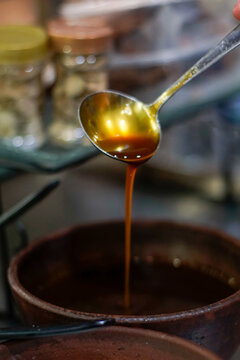 Close-up of liquid palm sugar (gula aren/kinca) being poured from a metal ladle into a rustic earthenware bowl. Warm amber syrup with glossy texture and artisanal kitchen background