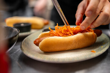 Close-up of hands preparing hot dog with shredded carrot topping on patterned plate in kitchen setting. Creative street food concept with healthy twist.