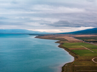 An aerial view shows a blue lake adjacent to green and brown farm fields under cloudy skies with distant mountains, highlighting rural and natural integration.