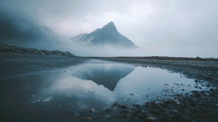 A mountain shrouded in mist reflects perfectly in a puddle on a dark rocky terrain