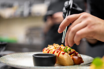 Close-up of a gourmet hot dog being garnished with chopsticks by a gloved hand. Served on a decorative plate with condiments and dipping sauce in a professional kitchen setting.