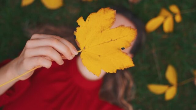 portrait happy young beauty woman lying on green grass view from above hand holding maple leaf hiding face. Cheerful girl enjoying life autumn nature yellow orange color fallen leaves. hello october