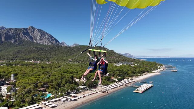 Two people parasailing over a tropical beach resort with turquoise sea, piers, green forest, and towering mountains under a bright blue sky.
