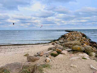 Kites flying above calm sea near sandy beach and stone pier under cloudy blue sky