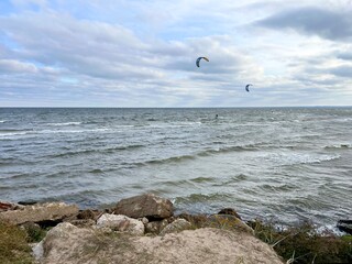 Kitesurfers gliding over sea waves near rocky shore under cloudy sky, coastal water sport scene