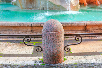 Stone bollard with rounded top and decorative iron supports, set against backdrop of splashing fountain with turquoise water. Urban setting with cobblestone pavement and part of larger water feature
