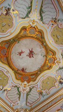 Close-up vertical shot of an exquisite Rococo or late Baroque ceiling fresco featuring three flying cherubs or putti in a central oval frame, surrounded by intricate gold leaf stucco work and delicate