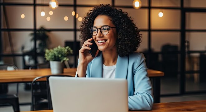 Smiling woman in a blue blazer talking on a smartphone with a laptop in front of her