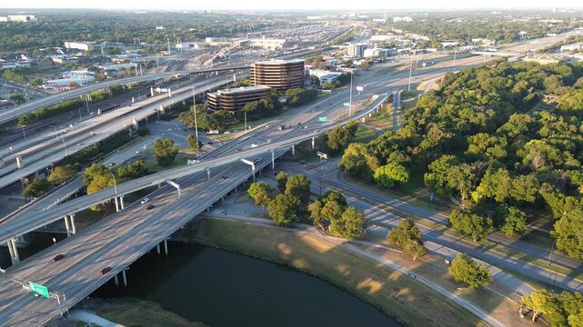 Lush urban park and tree canopy border the Trinity River, stacked highway ramps curving toward Fort Worth high-rise office buildings. Flyover blends mobility, green space, architectural density