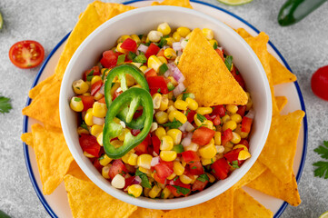 Corn salsa in a bowl with lime, jalapeno pepper, fresh herbs and tortilla chips on a concrete background. Mexican cuisine. Healthy food.