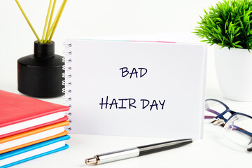 A notepad on a bright desk reads bad hair day. Near it are books, a pen, glasses, a plant, and a reed diffuser in a black vase, suggesting morning routines or study