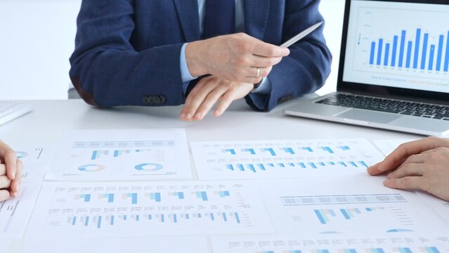 Businessman professional pointing laptop screen showing bar graphs, calculator on table while team collaborates on financial analysis and planning. Business people concept