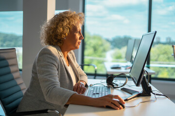 Mature businesswoman focused on her work, sitting at a desk with a computer and headphones, processing data in a modern office with large windows overlooking a green landscape