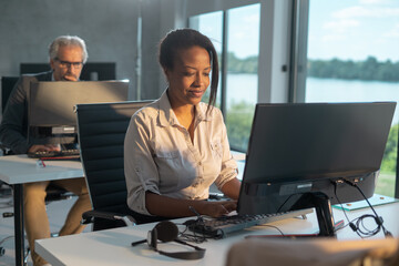 Young black businesswoman smiling and typing on computer keyboard at her desk, with a senior colleague working in the background, showing collaboration and contemporary workspace