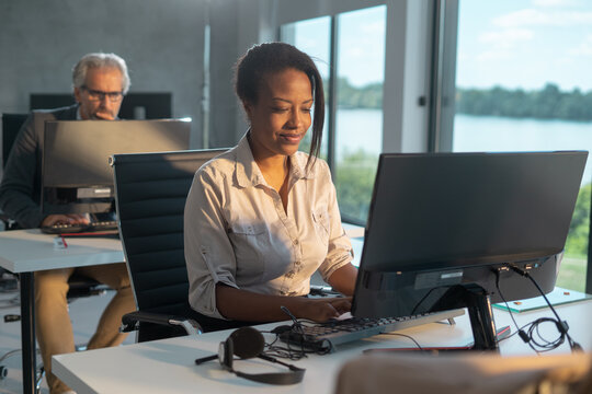 Young black businesswoman smiling and typing on computer keyboard at her desk, with a senior colleague working in the background, showing collaboration and contemporary workspace