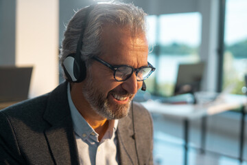Mature man wearing a headset and glasses, engaging in customer service, providing online support in a modern office environment, demonstrating professional communication and helpfulness