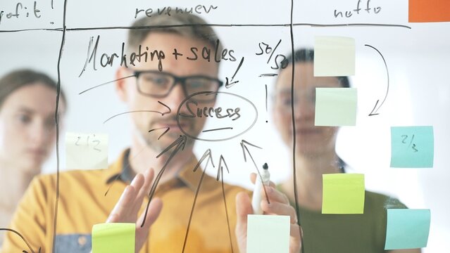Business people team brainstorming strategic goals, mapping innovative concepts on transparent whiteboard with colorful sticky notes and black marker in modern office