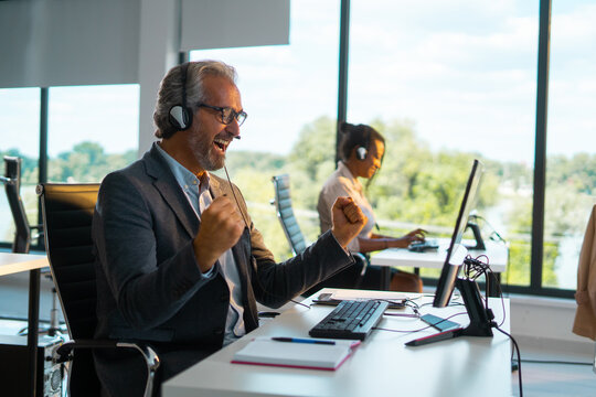 Senior businessman in headset pumps fist at his desk in a modern office, celebrating a successful sales call with a joyful, triumphant smile during a video or phone meeting - Powered by Adobe