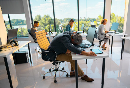Tired businessman feeling burnout and stress, sleeping at his desk during work after long hours in a modern office with colleagues working in the background