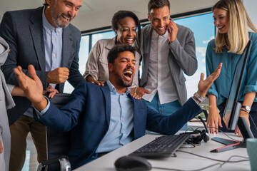 Diverse business people gathering around a computer. A man at the desk enthusiastically cheering and celebrating with hands raised. Colleagues smiling and showing support for their shared achievement