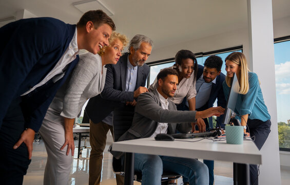 Diverse business team collaborating closely, gathering around a computer screen, reviewing data, and discussing new ideas and strategy in a bright contemporary office environment