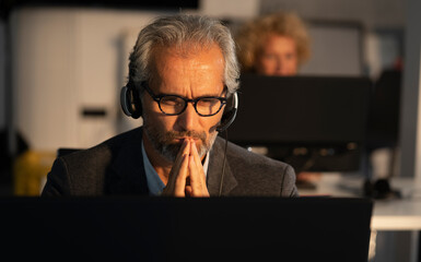 Mature professional with headset and glasses at a computer, hands clasped, lost in deep...