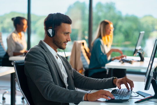 Male call center agent assisting customers, wearing headset and typing on computer keyboard in a bright, contemporary office environment with other operators in the background