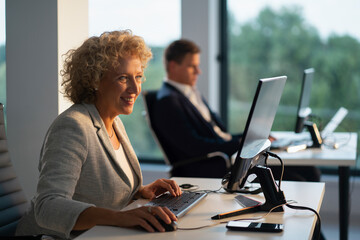 Smiling mid adult businesswoman working at her desk, typing on a keyboard and using a mouse while collaborating with a colleague in a bright, contemporary office environment