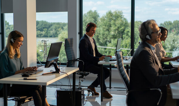 Diverse customer service team members wearing headsets and typing on computers, providing support to clients in a modern office with large windows overlooking a green landscape