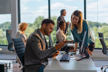Business colleagues working together in a contemporary open-plan office, a man writing on a clipboard while a woman stands by his desk, other diverse employees visible in the background