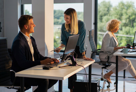 Business colleagues collaborating at an open-plan office workstation, smiling and discussing a project on a computer, demonstrating teamwork and professional interaction in a modern workspace