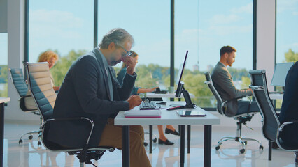 Overworked businessman rubbing his temples at a cluttered office desk, exhausted and stressed amid colleagues at computers, conveying burnout, workplace pressure and mental fatigue