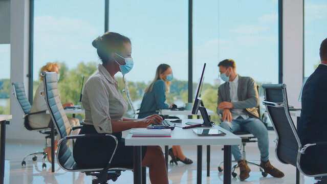 Diverse business team members wearing protective face masks, diligently working at their individual desks while maintaining social distancing in a modern, brightly lit open-plan office