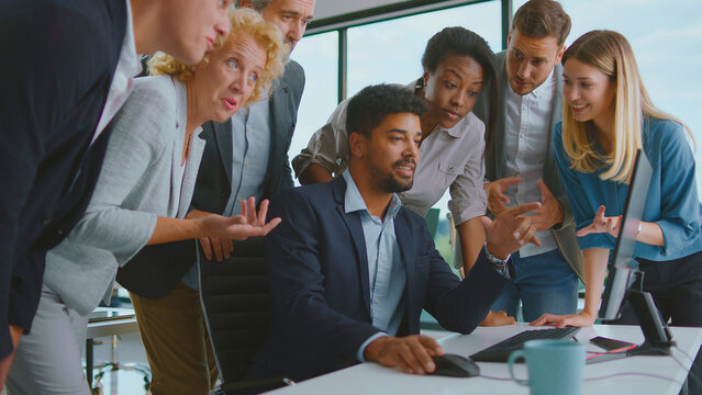 Diverse business professionals gathering around a computer screen in a modern office, working together and discussing ideas, showing teamwork and active collaboration