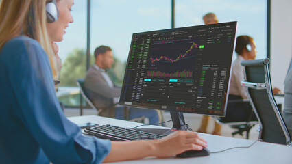 Businesswoman wearing headphones monitoring stock market data and analyzing financial charts on a computer screen, trading investments in a contemporary office