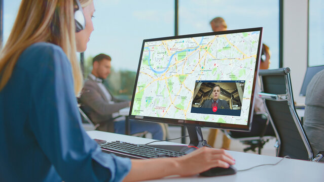 Woman wearing headset making a video call on a computer monitor displaying a map and a colleague while working at her desk in a modern open-plan office