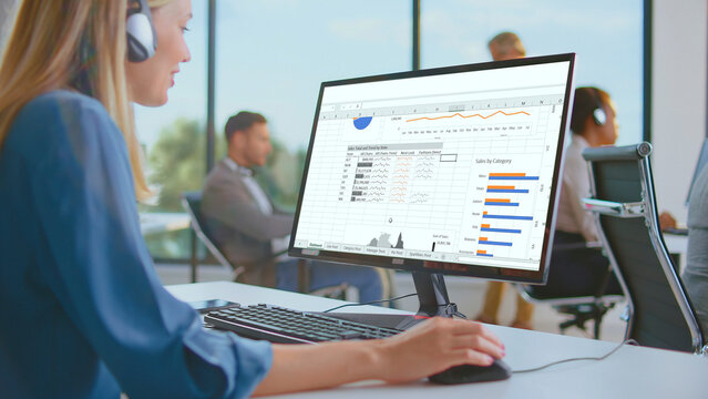 Woman wearing headphones working on a computer screen displaying spreadsheets and charts while analyzing financial data in a collaborative office environment - Powered by Adobe