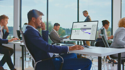 Professional diverse business team in a modern office, man smiling as he analyzes a project management gantt chart on his computer, focused on strategy, planning and collaboration