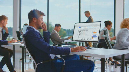 Professional man analyzing business data on computer spreadsheet, smiling in a collaborative open-plan office environment with colleagues working around him