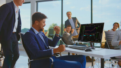Diverse team of happy stock traders in a modern office celebrating success and profit from a winning financial transaction while viewing a rising candlestick chart on a computer monitor