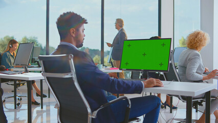 Young man using a computer with a green screen monitor in a modern corporate office, surrounded by diverse employees collaborating and managing tasks in a bright workplace