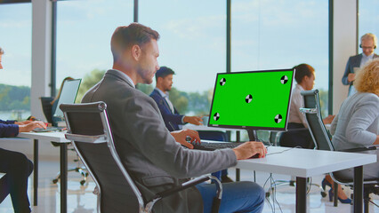 Employee working on a desktop computer with a customizable green screen display, collaborating with diverse colleagues in a modern open-plan corporate office
