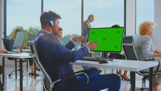 Customer support agent wearing a headset and pointing to a green screen monitor while working in a busy call center office providing diverse client assistance