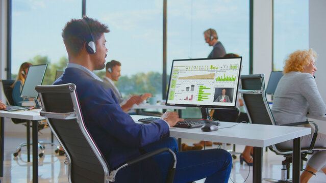 Customer service representative sitting at desk, wearing headset, analyzing financial charts and graphs on computer monitor while participating in a video call at a busy office