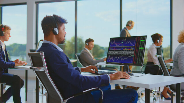 Diverse team of financial brokers wearing headsets, analyzing stock market charts and data on computers in a modern open-plan office during trading hours - Powered by Adobe
