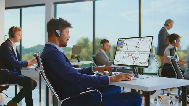 Diverse professional stock traders are working on computers with headphones, monitoring stock market charts and financial data in a bright, modern office environment